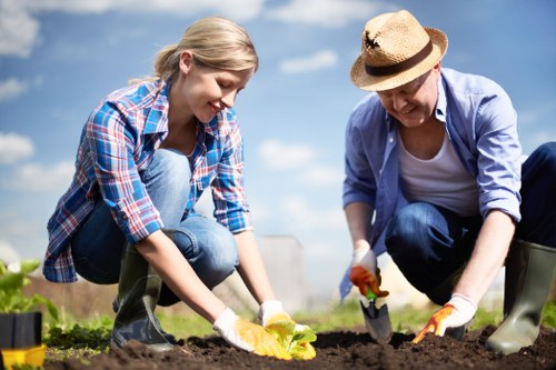 A woman and an older woman are working together in a garden, planting or tending to soil in a well-maintained outdoor space. The garden features a lush, green lawn with apparent dense grass in the foreground, bordered by flowering plants and shrubs along the edges. In the background, there are trees and a partly cloudy sky, suggesting a mild, sunny day. The woman and the older woman are dressed in casual gardening attire, including gloves and boots, actively engaged in soil preparation or planting. The scene highlights caring outdoor landscaping activities common in residential gardens around Crystal Palace, with focus on garden maintenance and planting tasks that enhance the outdoor environment's natural beauty.