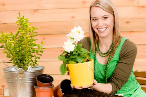The image depicts a smiling woman with shoulder-length blonde hair wearing a green polka-dot apron and a multi-strand green beaded necklace, seated in a garden setting with a wooden fence in the background. In front of her are various gardening pots, including a yellow pot she is holding containing a healthy, flowering plant with white blossoms and green foliage. To her left, there is a larger, silver metal pot with a young, vibrant green shrub, and nearby are empty terracotta pots. The surface appears to be a wooden garden table or workbench, supporting her gardening activities. The scene is outdoors with natural daylight, suggesting a calm, sunny day suitable for outdoor gardening and lawn care tasks. This image aligns with services offered by Lawn Mowing Crystal Palace, emphasizing garden plant care and pot planting in local outdoor spaces within Crystal Palace or the surrounding London area.