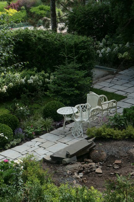 A small, neatly maintained garden scene featuring a paved patio area with a decorative wrought iron garden bench centered against a white building wall with a window and closed shutters. Surrounding t